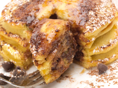 Stack of four golden chocolate laced pancakes dusted with cocoa and powdered sugar. Butter melting atop. Fork lifting first bite.
