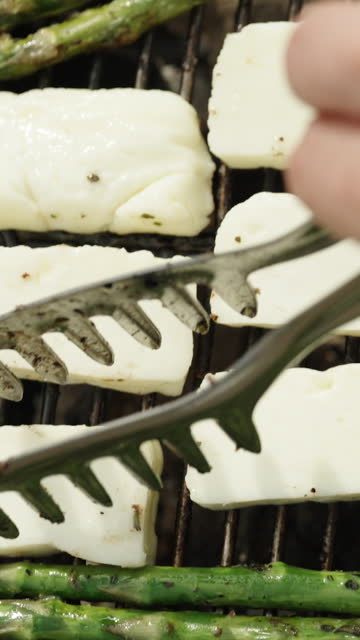 I am flipping the pieces of Haloumi cheese that are being grilled along with the asparagus on the charcoal grill. Vertical video.