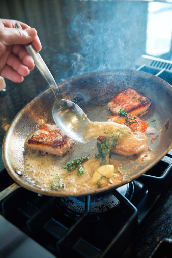 A chef's hand deftly bastes pieces of pan seared salmon with garlic and herbs in a fry pan.  Shallow dof, some motion blur