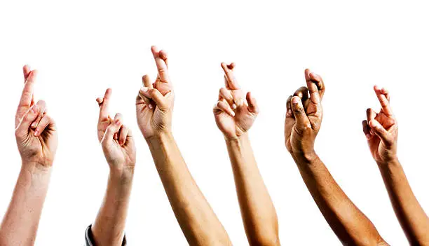Six mixed hands are raised, all with fingers crossed superstitiously, hopefully averting bad luck. Isolated on white. 