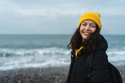 cheerful pretty female in yellow knitted hat and black jacket standing on seaside against ocean background and looking at camera