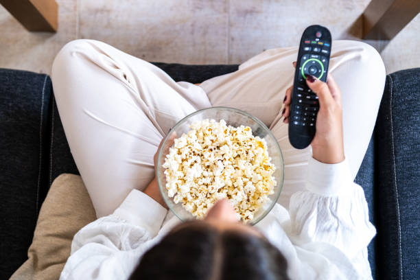 istock A young woman has prepared popcorn and searches for a program on TV with the remote control from her living room sofa. Top view. 1682572524