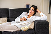 A young woman watches a movie or program on television while eating popcorn on the couch in her living room.