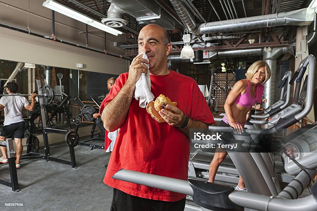 Enjoy your meal A man just can't resist a blueberry muffin in the middle of a workout session as his wife looks on with disgust, Gym Stock Photo