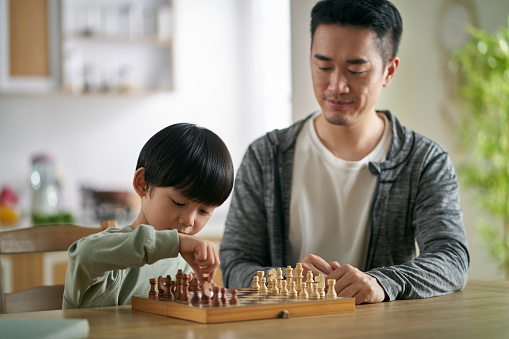 asian father and son sitting at kitchen table at home playing chess