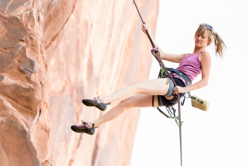 Young Woman Rock Climber at Wall Street near Moab Utah.  Athletic caucasian climber is rappelling down a cliff wearing crop pants and a lime green tank top, she has blond hair with a pony tail.  There is ample copy space in the image for titles and other design elements.