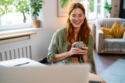 Young redhead Caucasian woman with vitiligo drinking coffee while having a video call via laptop