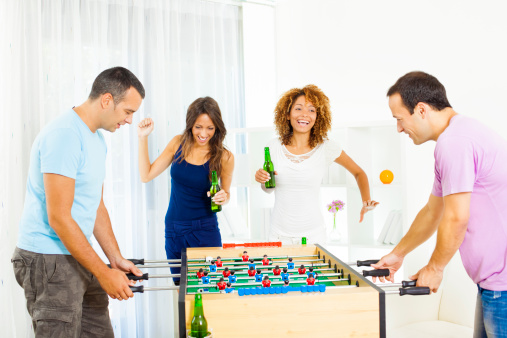 Two couples playing table footie at home and having fun. Men playing table footie and their girlfriends supporting them, jumping and shouting.