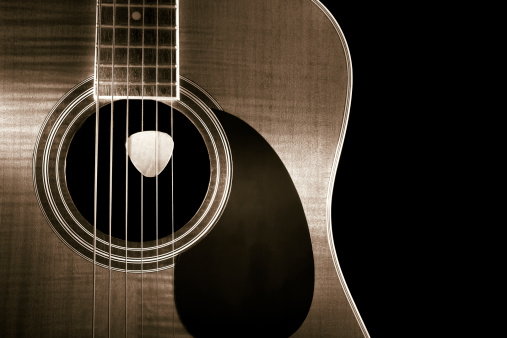 Toned BW photo of maple wood acoustic guitarwith pick showing wood grain, bridge and strings.