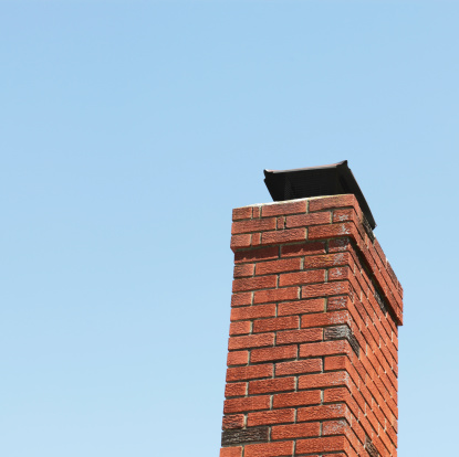 A red brick chimney - with a capped metal grate on top to prevent rain, birds and pests from entering - against a blue sky. Square composition.