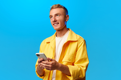 Portrait of young handsome smiling guy, student dressed in bright fashion outfit holding phone isolated on blue background. Concept of human emotions, youth, beauty, study and working online.