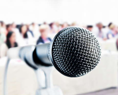 Close up on a vocal microphone in front of an out-of-focus audience at a meeting, seminar or social occasion.