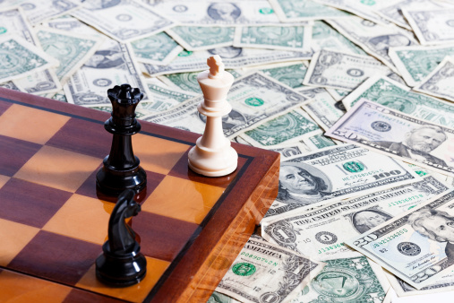 A chessboard with chessmen positioned for checkmate on it rests on top of a pile of American dollars. 