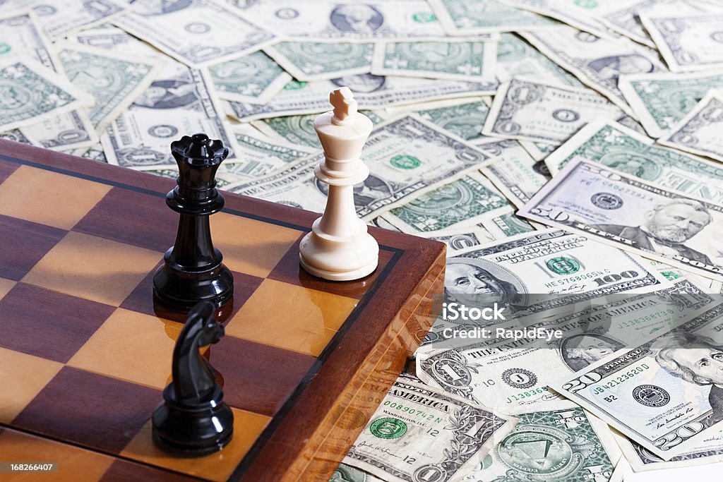 Checkmate! Chessmen and chessboard resting on pile of US currency A chessboard with chessmen positioned for checkmate on it rests on top of a pile of American dollars.  King - Chess Piece Stock Photo