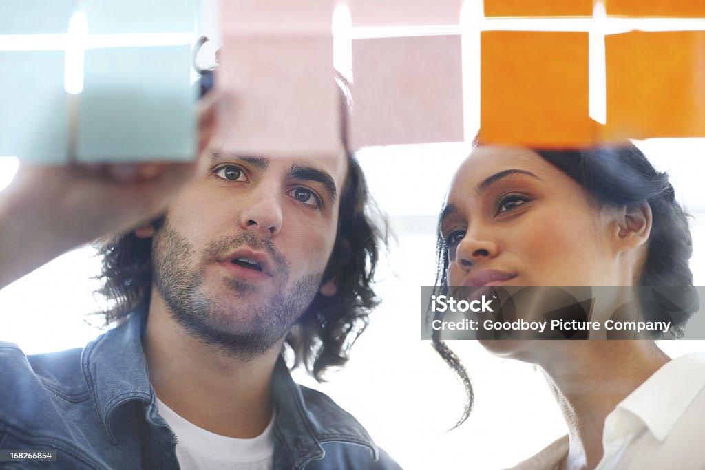 Sharing his creative opinions Two young colleagues surveying some notes stuck on a glass wall Adult Stock Photo