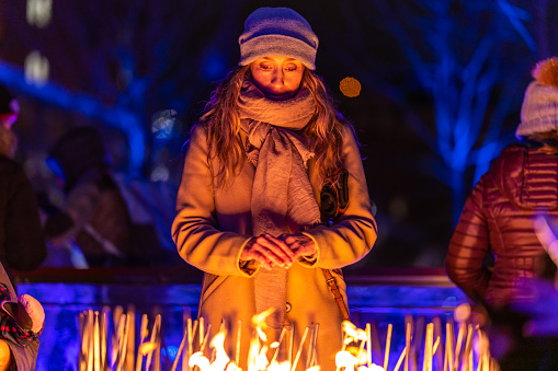 A Portuguese woman enjoying the Montreal Christmas market at night and warming her hands near a fire pit.