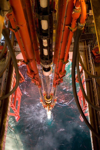 offshore oil rig riser pipe view to sea through moonpool viewed from height. Main pipe contains drilling fluids and drill pipe and connects to the seabed wellhead equipment. Mud line hoses and hydraulic tensioners also seen. Promotes careers, industry, oil, environment, energy and financial issues. Remote workplace in isolated environment with advanced technology.