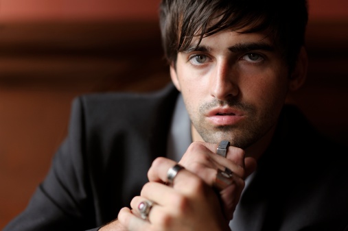 Portrait of a handsome, fashionable young man wearing a tuxedo jacket over a black t-shirt and chunky metal rings on his fingers. Shallow depth of field with sharp focus on the model's left eye.