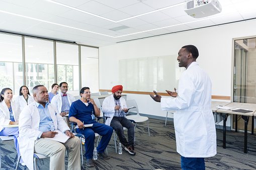 In the hospital meeting room, the young adult male doctor teaches a seminar for the diverse group of medical professionals