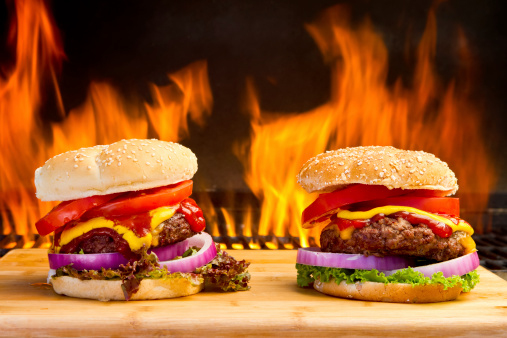 Two giant cheeseburger with all the toppings, tomato and onion, cheese, ketchup and mustard on a bamboo cutting board with big flames leaping in the background.  Sandwiches are placed at the lower half of the image making ample copy space for titles or any other design elements.