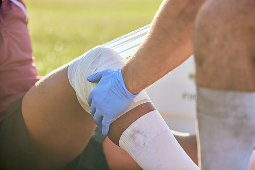 First aid, sport injury bandage and hands on knee with soccer accident, fitness and massage on a field. Training, workout and physical therapy of leg pain at game with emergency from exercise