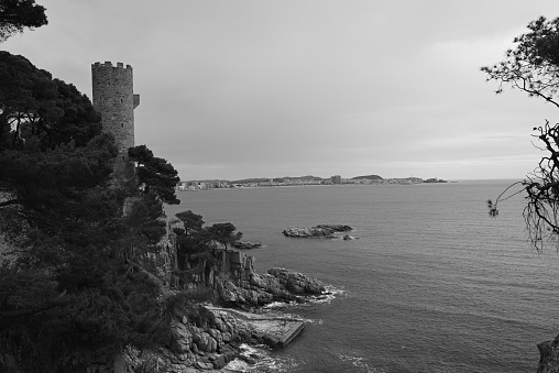A grayscale view of a rocky hillside with a tower overlooking the water in Costa Brava