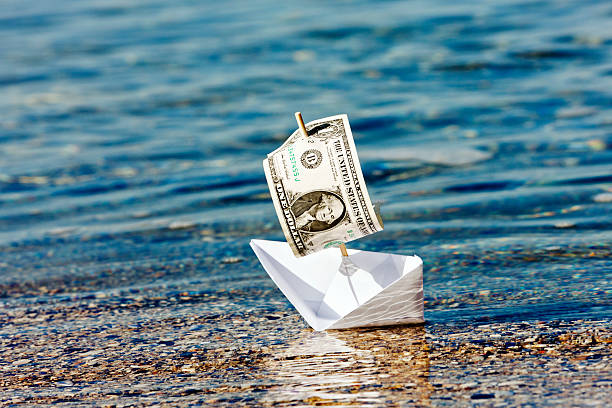 Paper boat with $1 sail stuck in shallows: money problems? A paper boat with a $1 bill for a sail sets off to sea, but seems stuck in the shallows. Symbolizes a poor investment, sinking dollar, or simply starting out financially. sports betting exchange stock pictures, royalty-free photos & images
