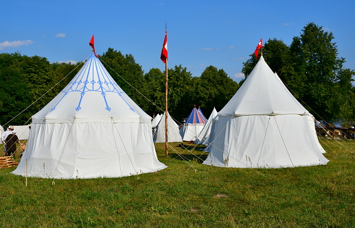 A close up on a temporary camp made out of white and colorful cloth tents with some medieval equipment visible spotted during a knight themed fair or festival organized in summer on a vast field