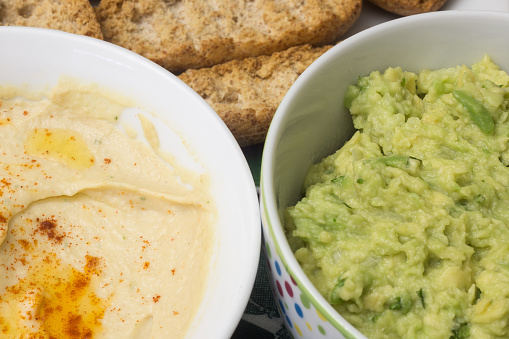 A very close-up shot of two bowls of dip, one with hummus and one with guacamole, both made with fresh ingredients. The breadsticks and toast are out of focus. A tempting and nutritious snack.
