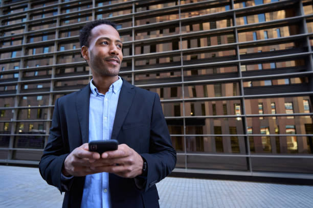 istock Young Latin professional male looking curious to side using smart phone center building background 1682898533