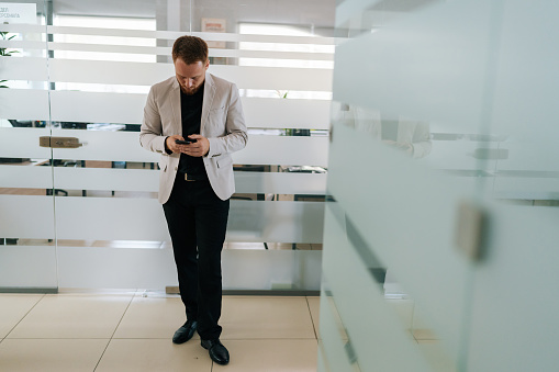 Full length portrait of busy businessman in suit using mobile phone standing in light hallway corridor of modern business center. Professional business man holding smartphone, working on cellphone.