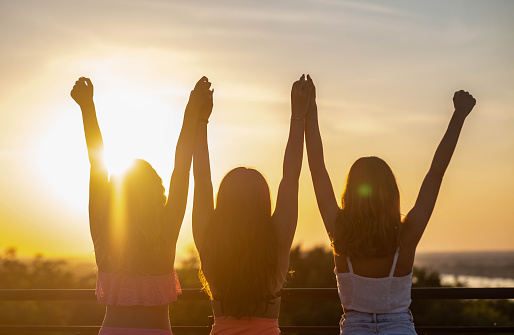 three girls with raised arms standing outside at sunset side by side