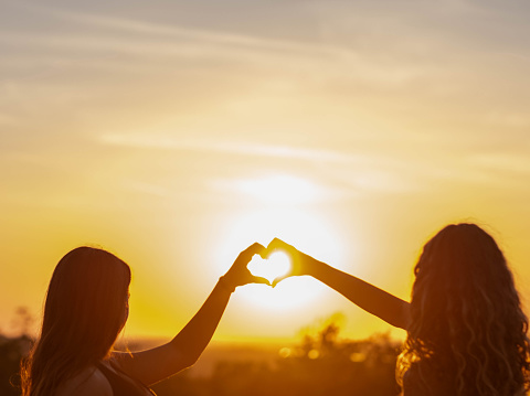 two girls are standing outside watching the sunset and making a heart shape with their hands