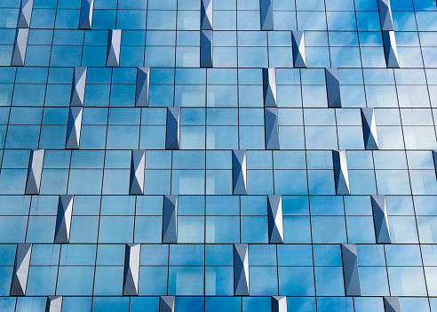 Blue glass and pyramids pattern on a commercial building façade - curtain walls form the outer shell of a building and stands as a separate shell in front of the actual supporting structure. They usually runs across the floors. Since the wall only supports its own weight and no other static loads from the building, it can be designed as a lightweight structure. The curtain wall is attached or suspended from the building's supporting structure using a substructure. The façade spanning several floors usually has a frame construction made of steel or aluminum profiles, which is largely filled with glass or other flat filling elements.