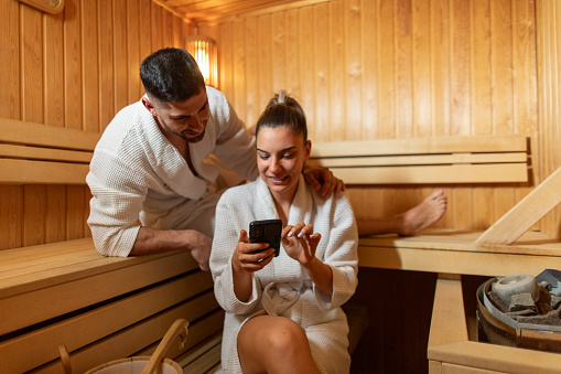 Beautiful young adult couple sitting in sauna and enjoying their time together. They are using mobile phone, wearing bathrobes and they are looking happy