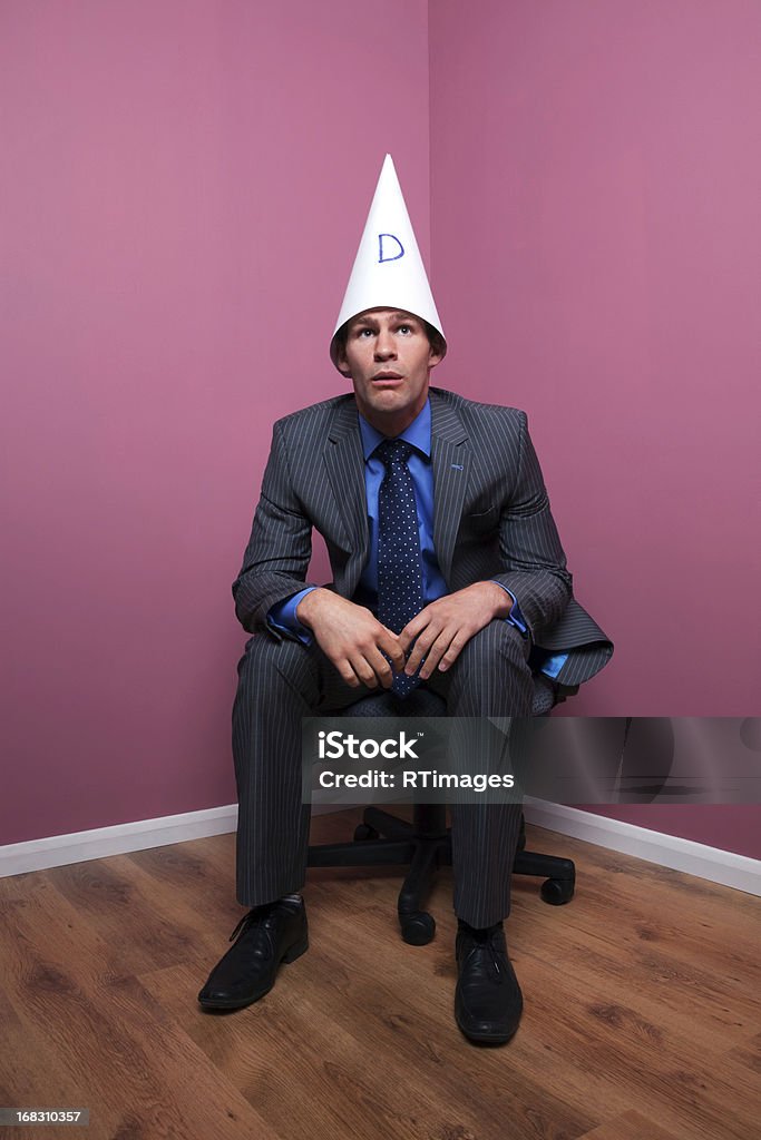 Biznesmen sat w rogu na sobie kapelusz dunce - Zbiór zdjęć royalty-free (Czapka matołka)