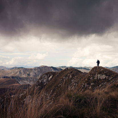 Mountaineer standing on the top of mountain peak and looking over endless massif area, dark sky and clouds above him.