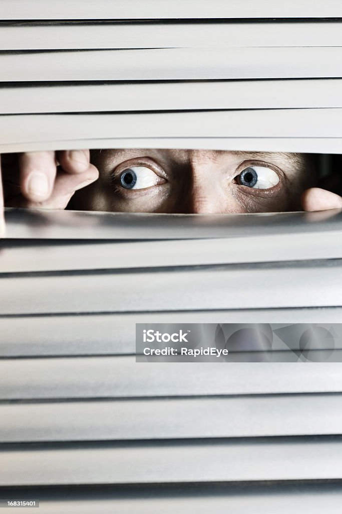 Wide eyed, worried man looks through venetian blinds Wide-eyed  and worried man peeps out through the slats of a closed  venetian blind, looking fearful.   Fear Stock Photo