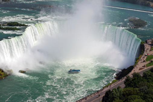 An aerial view of Niagara Falls. The lakes feed into the massive falls with white water rushing over the side. The Maiden of the Mist boat is cruising around the bottom of the falls. There is a pedestrian walkway and observation path overlooking the falls.