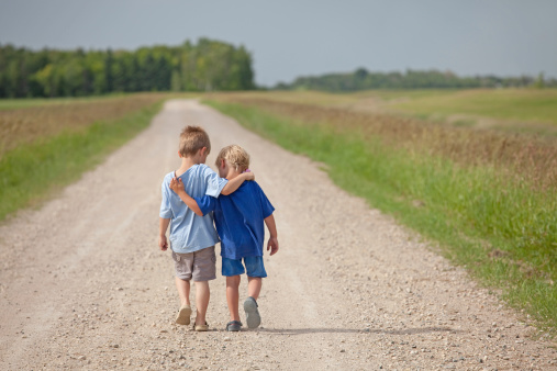 Two young boys walking arm in arm down a rural country gravel road. Prairie scene. Elementary aged caucasian children. Horizontal color image. Additional themes include friendship, bonding, boys, embracing, hugging, love, relationships, brothers, kindergarten, preschool, talking, care, best friends, and summer. 