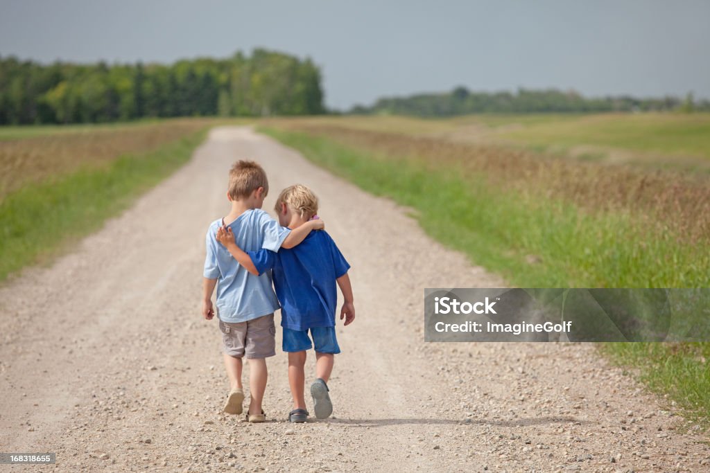 Two Caucasian Boys Walking Down a Country Road Two young boys walking arm in arm down a rural country gravel road. Prairie scene. Elementary aged caucasian children. Horizontal color image. Additional themes include friendship, bonding, boys, embracing, hugging, love, relationships, brothers, kindergarten, preschool, talking, care, best friends, and summer.  Child Stock Photo