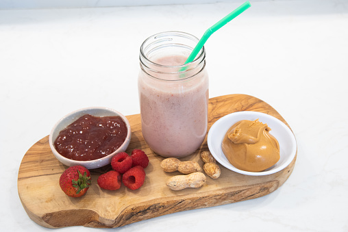 Peanut butter and jelly smoothie in glass with straw on cutting board with ingredients