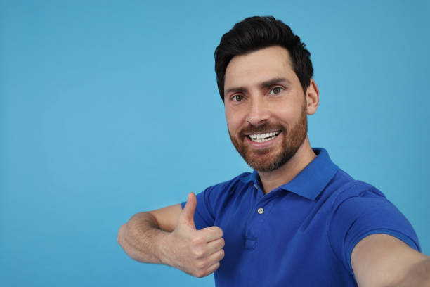 istock Emotional man taking selfie and showing thumbs up on light blue background, space for text 1683247559