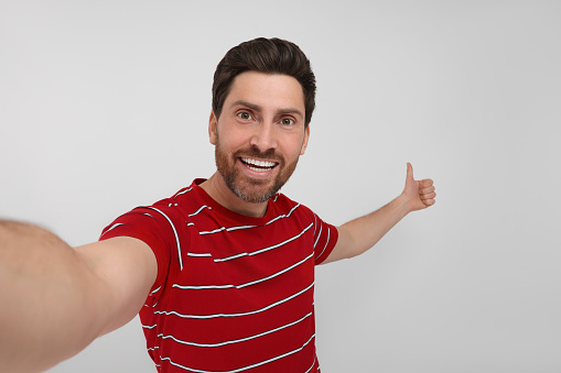 Smiling man taking selfie and showing thumbs up on white background