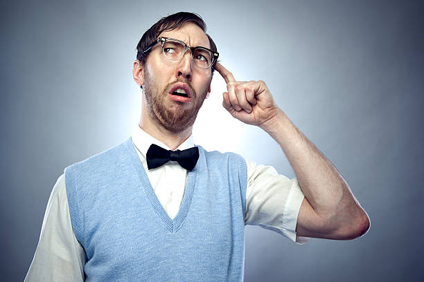 Concentrating Nerd Student Scratching Head Nerdy IT young man wearing a bow tie, blue sweater vest, and horn rimmed glasses makes a confused, goofy face while trying hard to think of something important. Horizontal on gray / blue background with copy space. nerd stock pictures, royalty-free photos & images