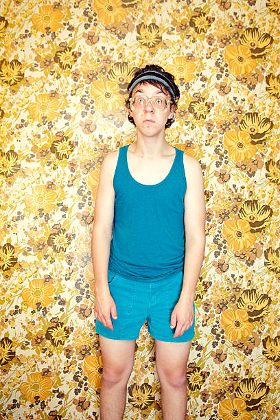 Nerd Young Man Looking Embarrassed A goofy, nerdy teenager with tank top, corduroy shorts, sweatband, and glasses, looks lonely and scared ad he stands in a vintage yellow room.  Cool retro floral wallpaper on the walls.  Vertical with copy space. nerd teenager stock pictures, royalty-free photos & images