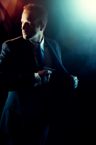 A confident, well-dressed male agent reaches into his coat and pulls out a handgun while looking over his shoulder, partially hidden in darkness. Bright street/stage lights glow behind him creating a high contrast profile. Vertical with copy space.