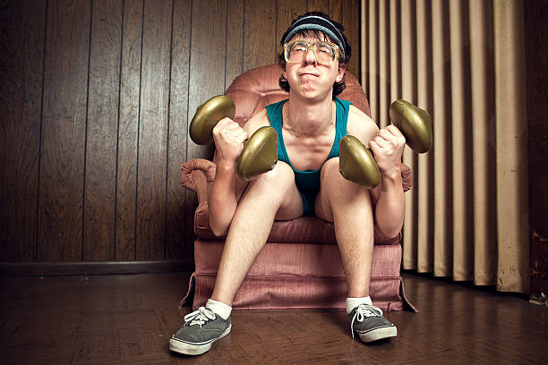 Nerd Young Man Exercising with Weights A goofy, nerdy teenager with tank top, corduroy shorts, sweatband, and glasses, strains to lift his dumbbell weights in a vintage basement, complete with linoleum and wood paneling on  the walls.  He sits in a plush pink armchair. Horizontal with copy space. weakness photos stock pictures, royalty-free photos & images