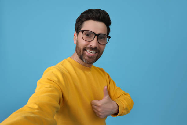 istock Smiling man taking selfie and showing thumbs up on light blue background 1683250739