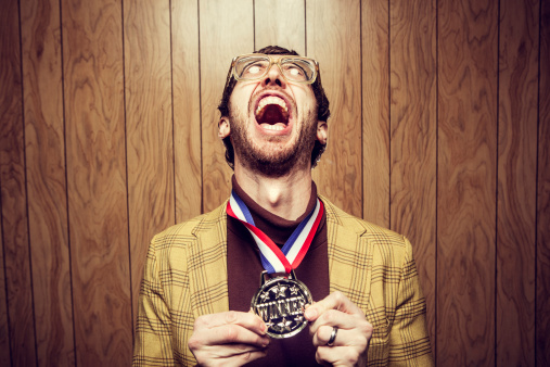 A nerd in a turtleneck and ugly yellow blazer with oversized glasses, bad hair, and a funny face celebrates proudly in front of retro wood paneling with his gold medal prize for winning.  Horizontal with copy space.